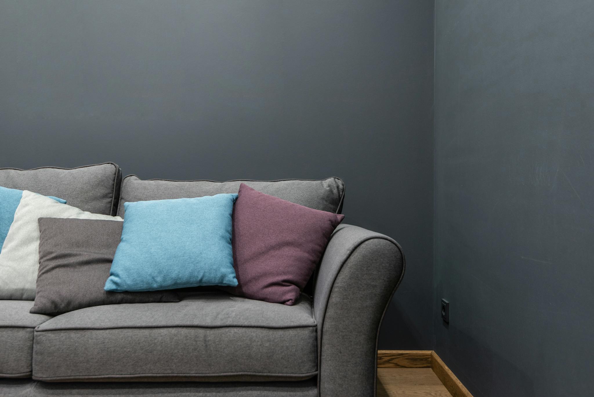 Part of soft modern gray sofa with armrest and abundance of bright pillows placed near wall in light room at home